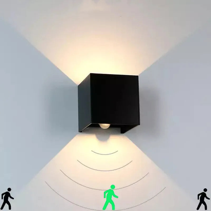 Small square aluminium wall lamp in black, mounted on a corridor wall, emitting a warm white light.