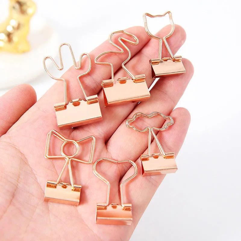 Small gold metal office clip with a long tail handle, shown in decorative bow, cactus, cat, flamingo, and love shapes.