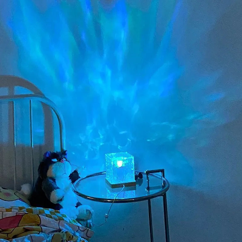 LED water ripple night light with 16 colours, USB cable, and remote control on a bedside table