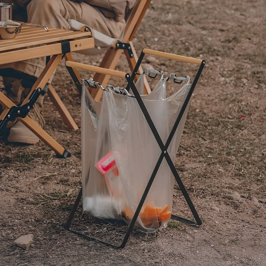 Black outdoor folding trash rack with wooden handle holding a white bin bag open.